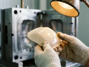 Gehandschoende handen inspecteren een glanzend kunststof spuitgietonderdeel onder precisielicht, met een stalen matrijs op de achtergrond.